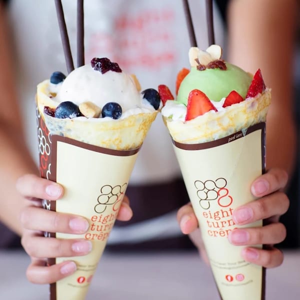 Two hands holding Eight Turn Crepe desserts, one with vanilla and blueberries, the other with green tea and strawberries.