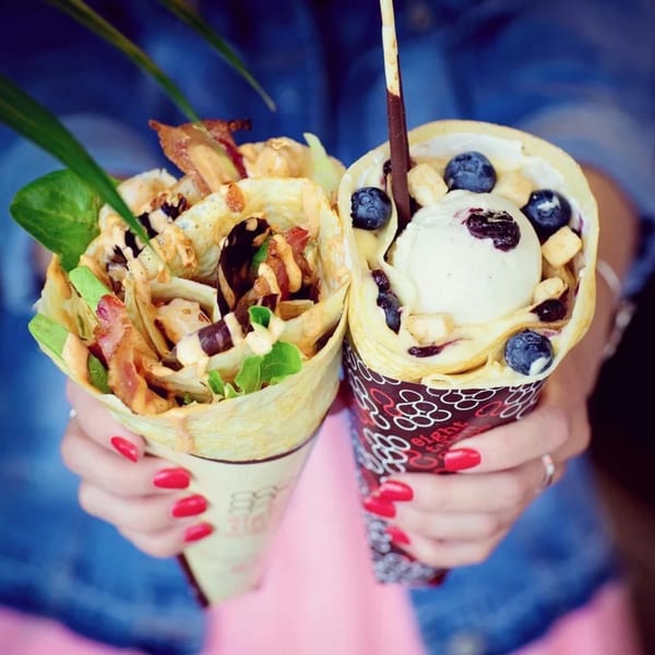 Hands holding two Eight Turn Crepes: one savory with bacon and greens, one sweet with ice cream and blueberries.