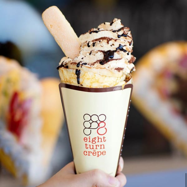 A hand holds an Eight Turn Crepe with whipped cream, chocolate drizzle, cocoa powder, and a ladyfinger.
